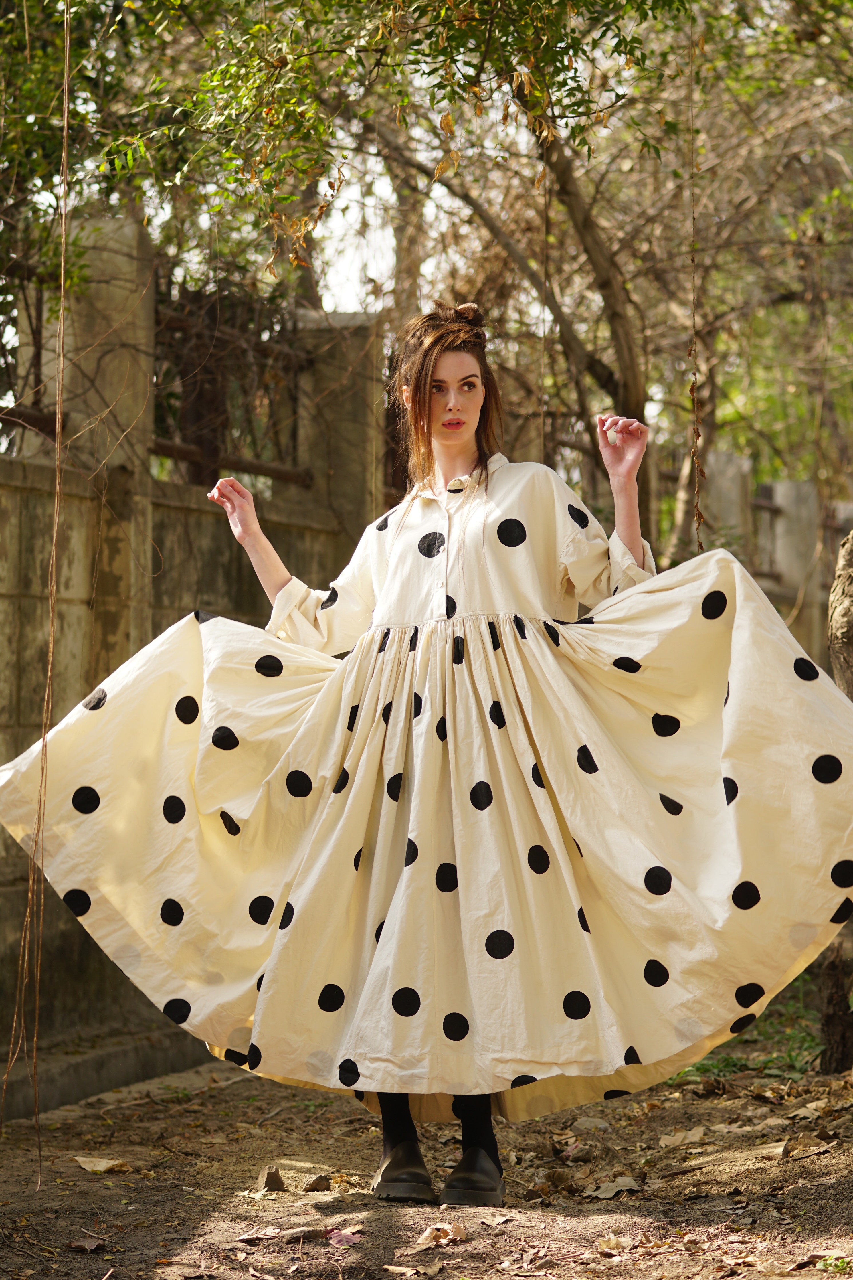 A MegbyDesign model is wearing a long cream dress with large black spots. The dress has side pockets, double stitching and a tiny collar to accent and give detail to the bodice.