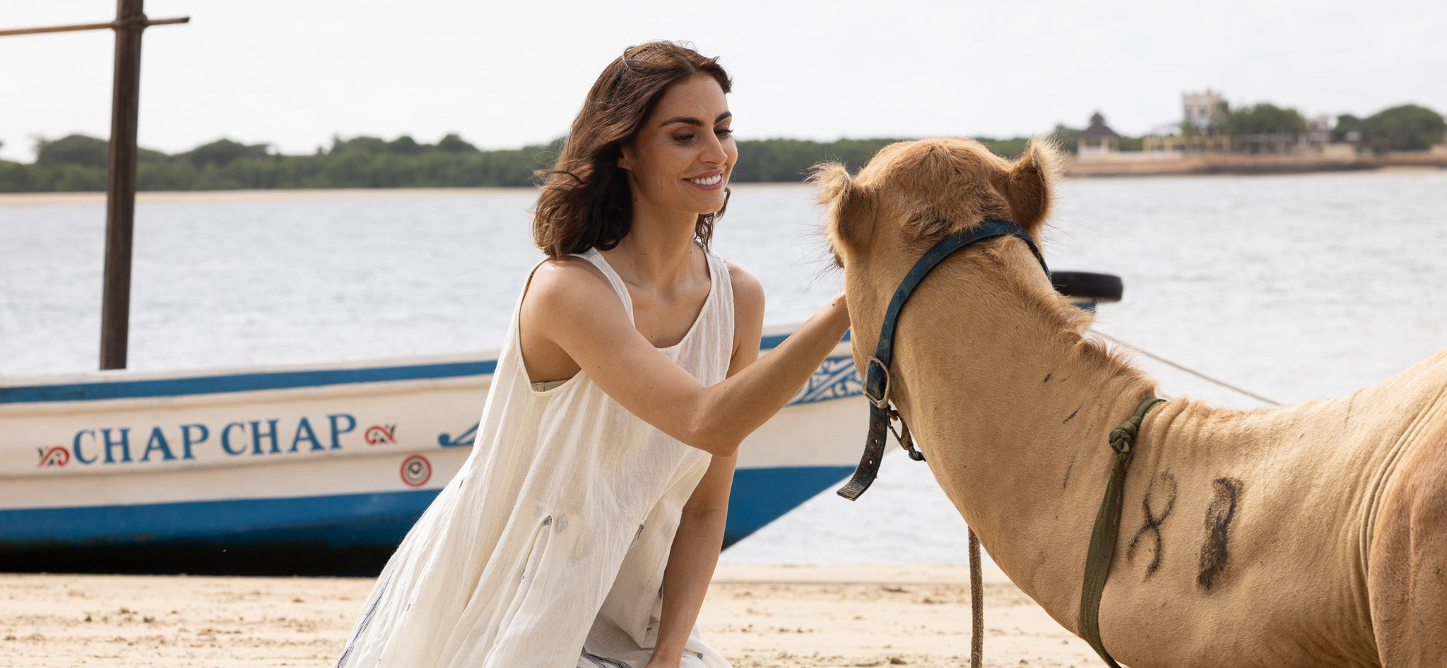 Woman in a white dress petting a camel on a beach with a boat in the background.
