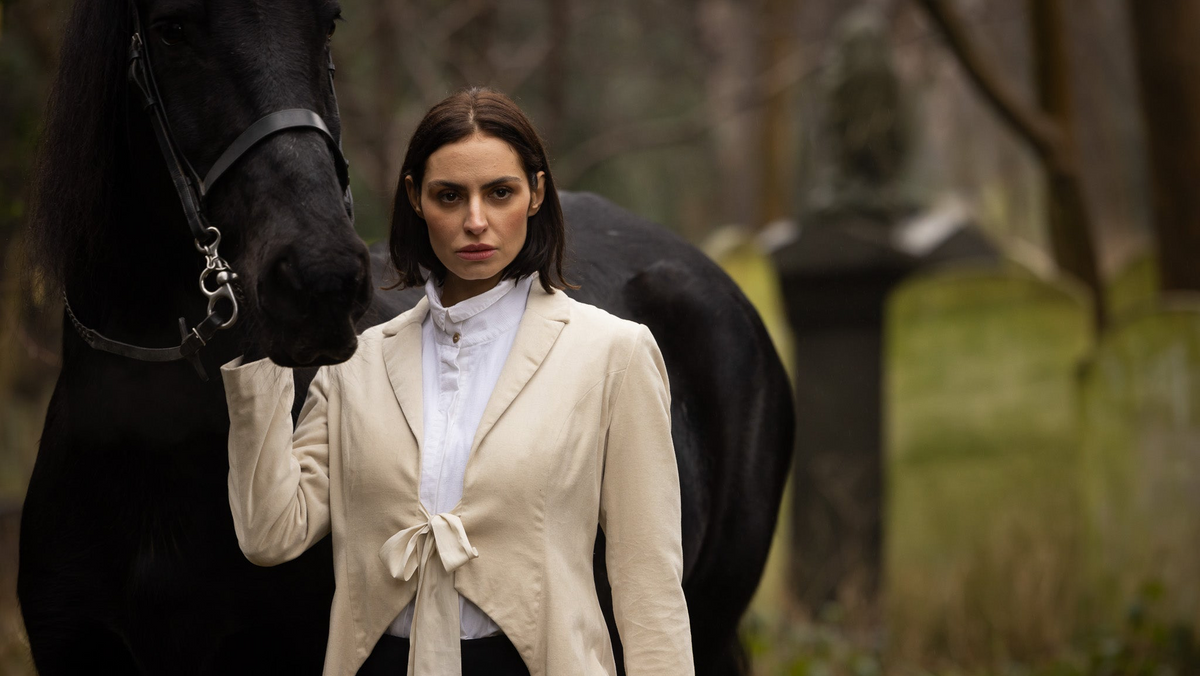 A woman stands outdoors beside a large black horse, wearing a cream tied-front coat over a white high-neck top and dark bottoms. The background is a softly blurred wooded setting with a stone monument.