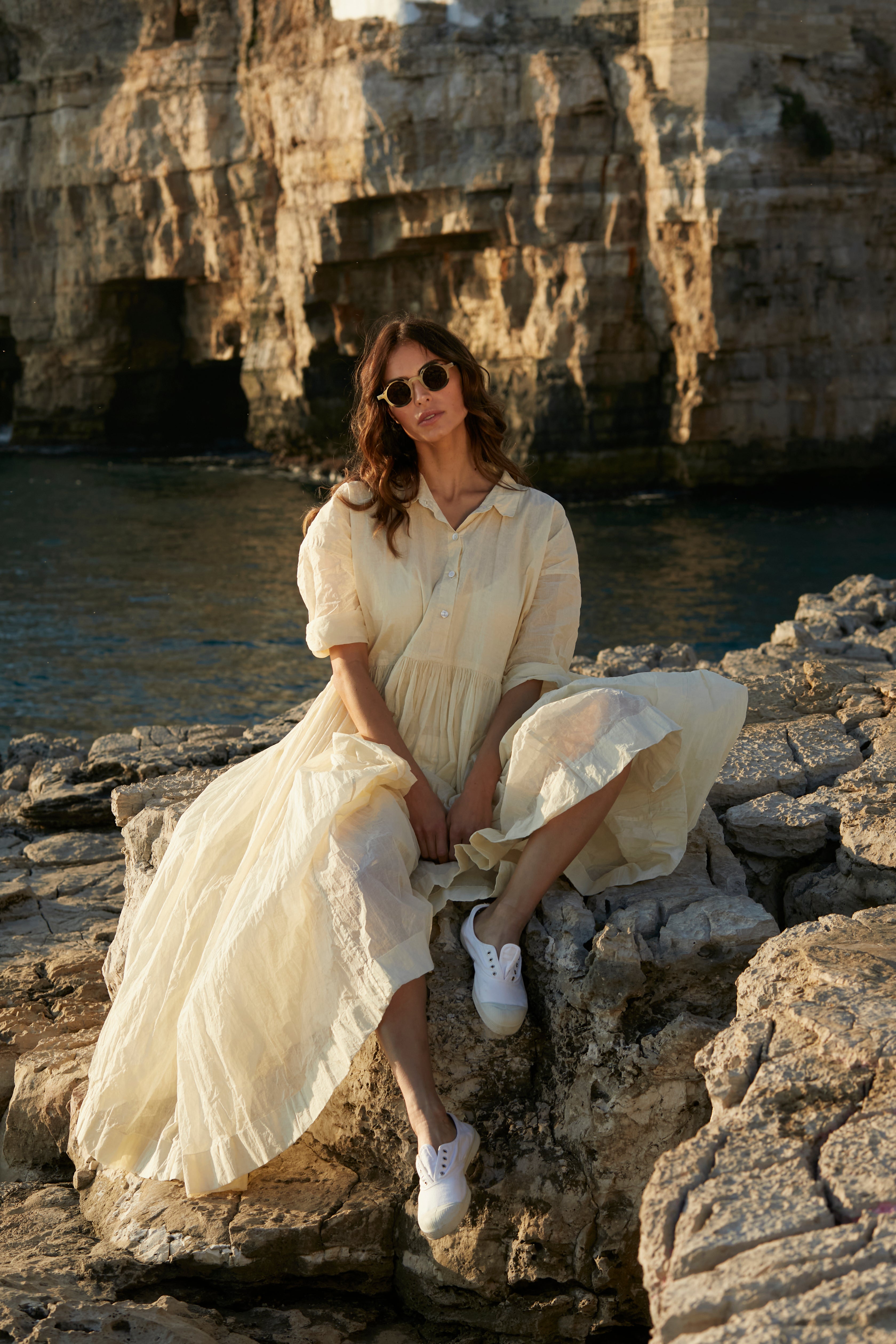 A MegbyDesign model is wearing an ecru edith dress made from a gorgeous organdy material. The model is sitting on a rock by the sea.