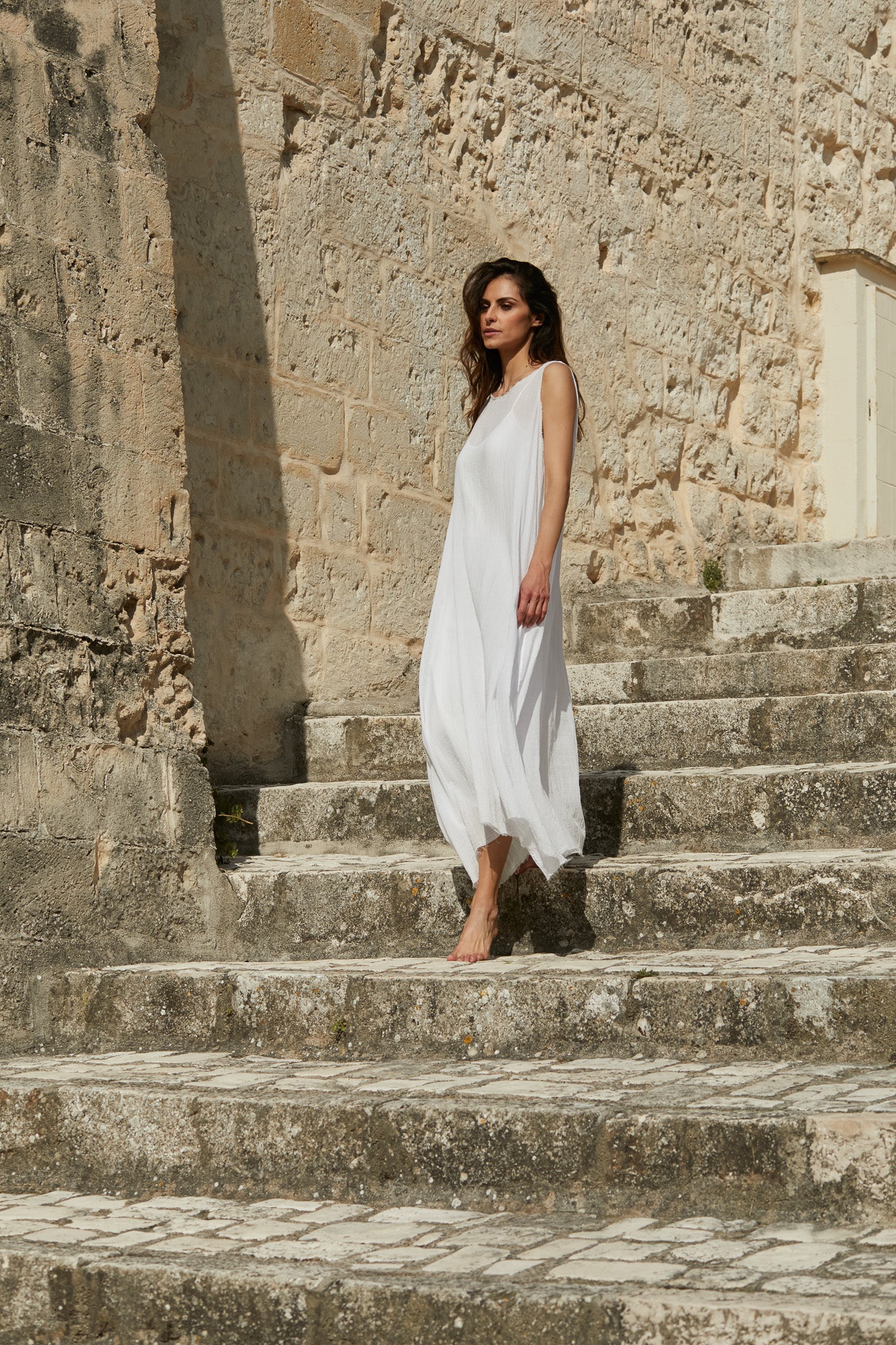 A MegbyDesign model is wearing a white gauze cotton angelica dress which is made from a beautifully lightweight gauze cotton. The model is bare foot and walking down a set of cobblestone steps