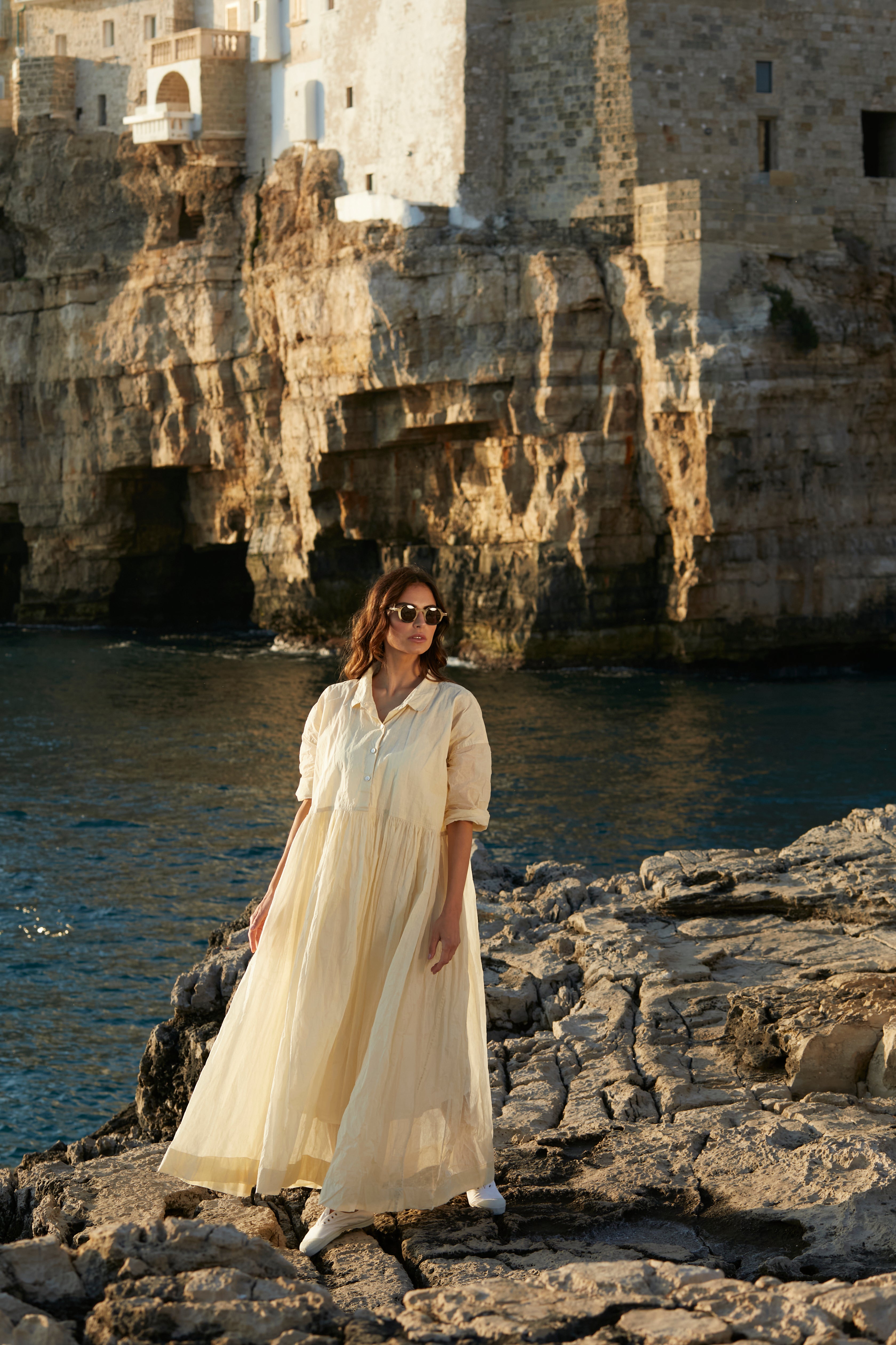 A MegbyDesign model is wearing a ecru edith dress made made from a gorgeous organdy material. The model is standing on a rocky shore line.