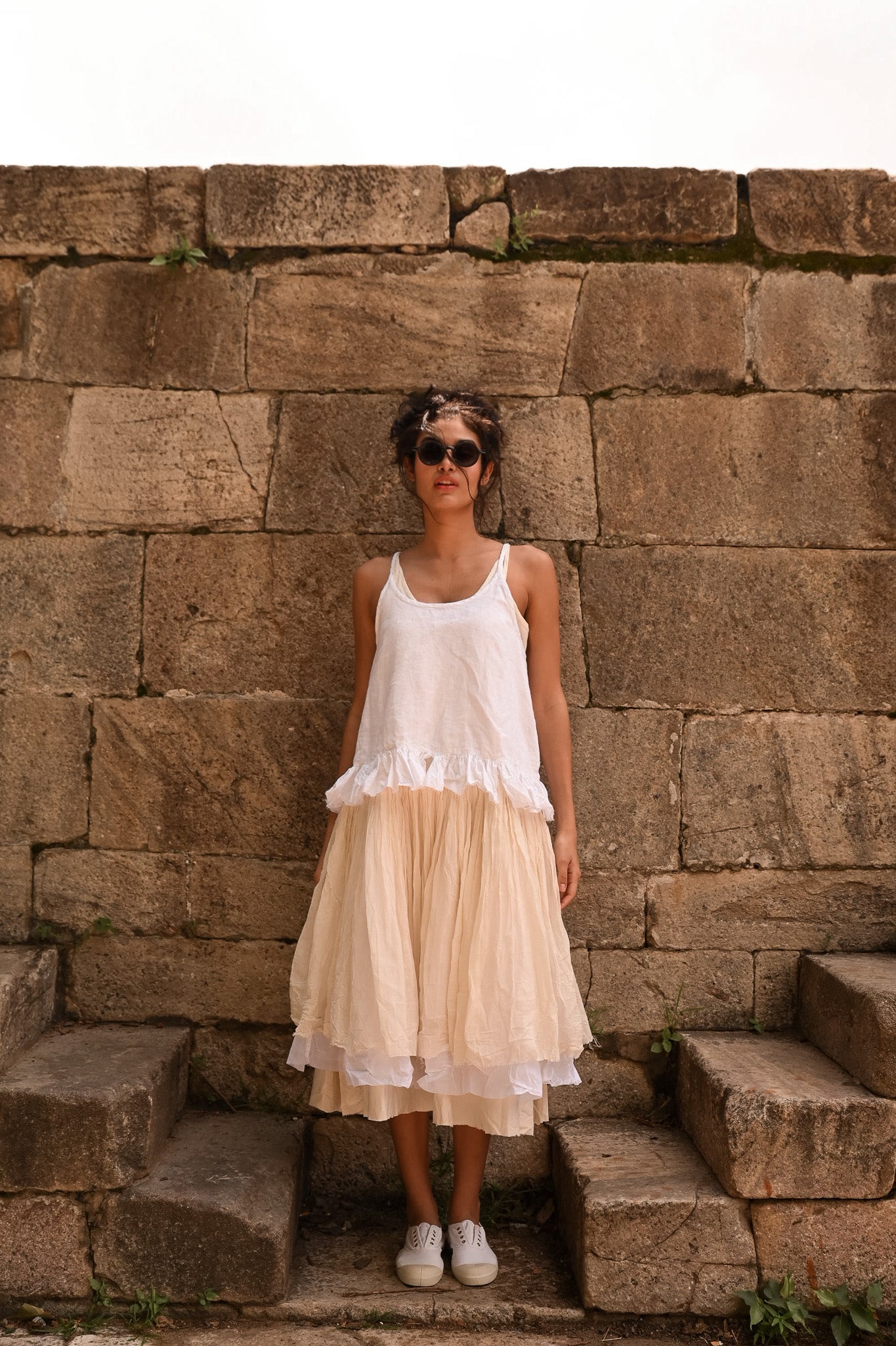 A MegbyDesign model is wearing a white elisa cami made from a lightweight cotton material and the straps are adjustable. It is a full body photo and the model is standing in from of a block stone wall.