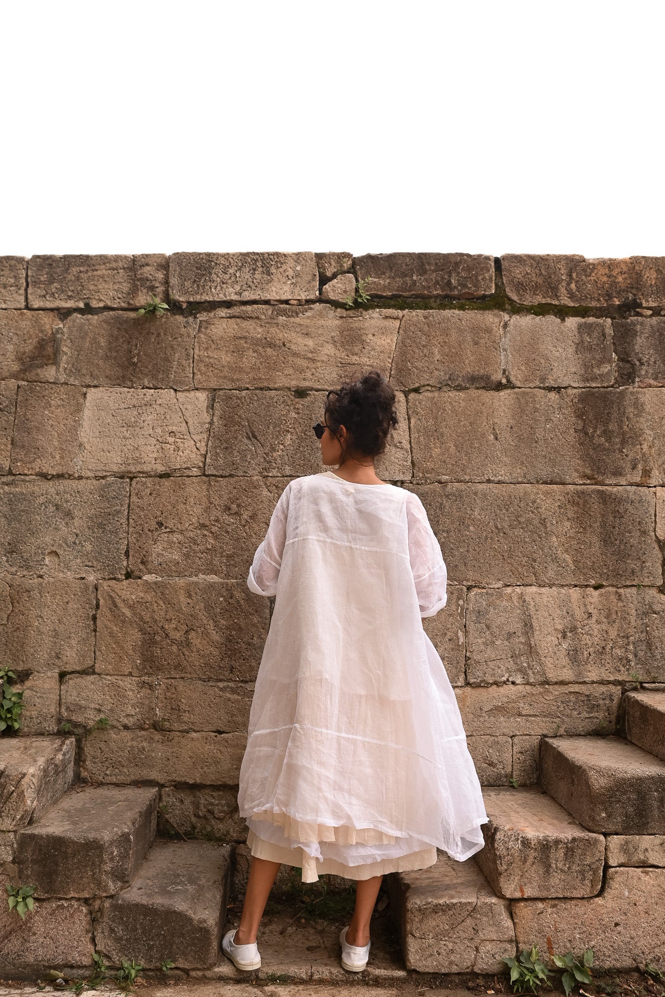 A MegbyDesign model is wearing a white prachi coat made from a crinkly cotton organdy material, she is standing but facing away from the camera.