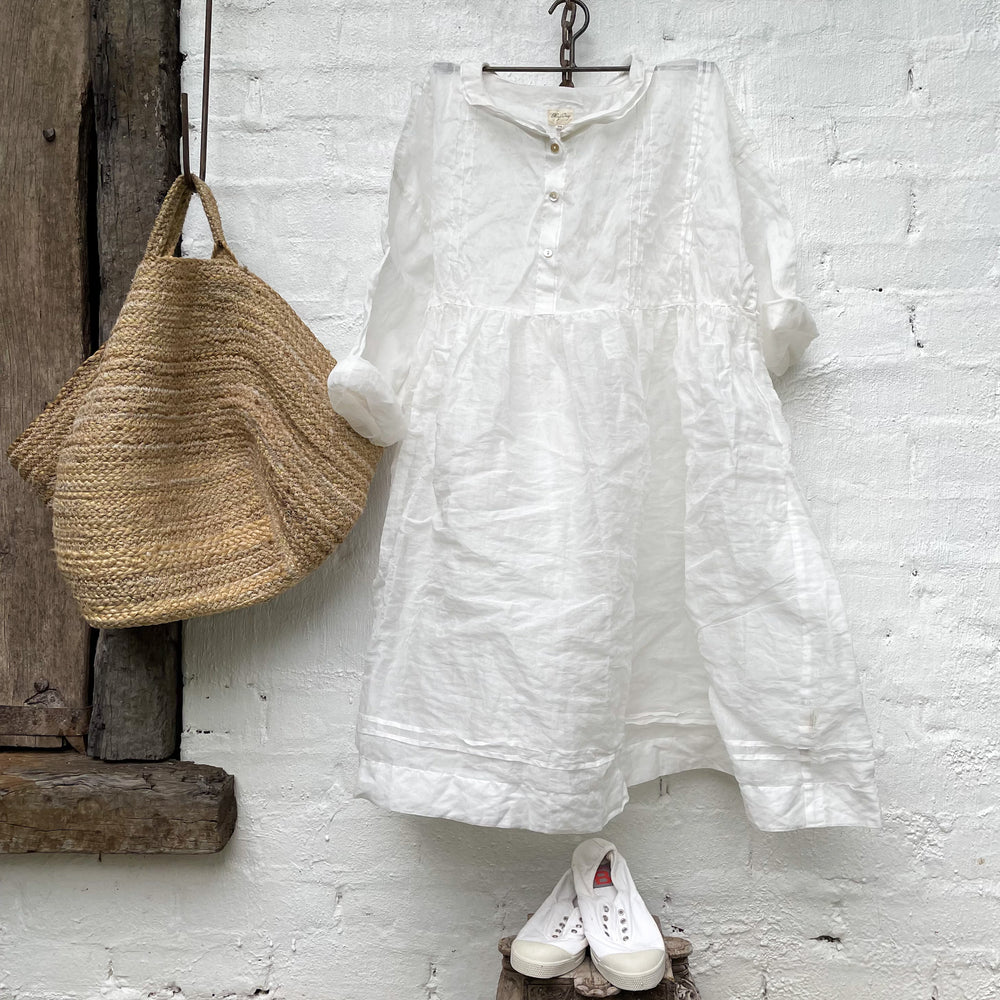 MegbyDesign cotton organdy dress hanging on a hanger in front of a white wall, a raffia bag hanging next to it, and white shoes placed on a stool. 